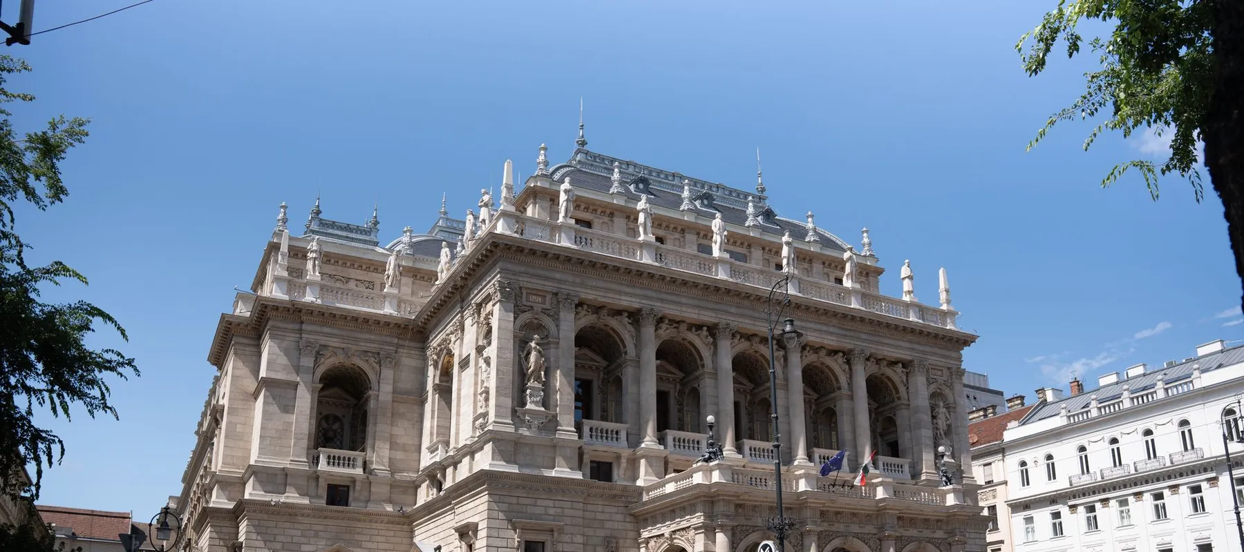 La fachada de la Ópera Estatal Húngara en Andrássy út bajo un cielo azul, con su línea de tejado flanqueada por estatuas