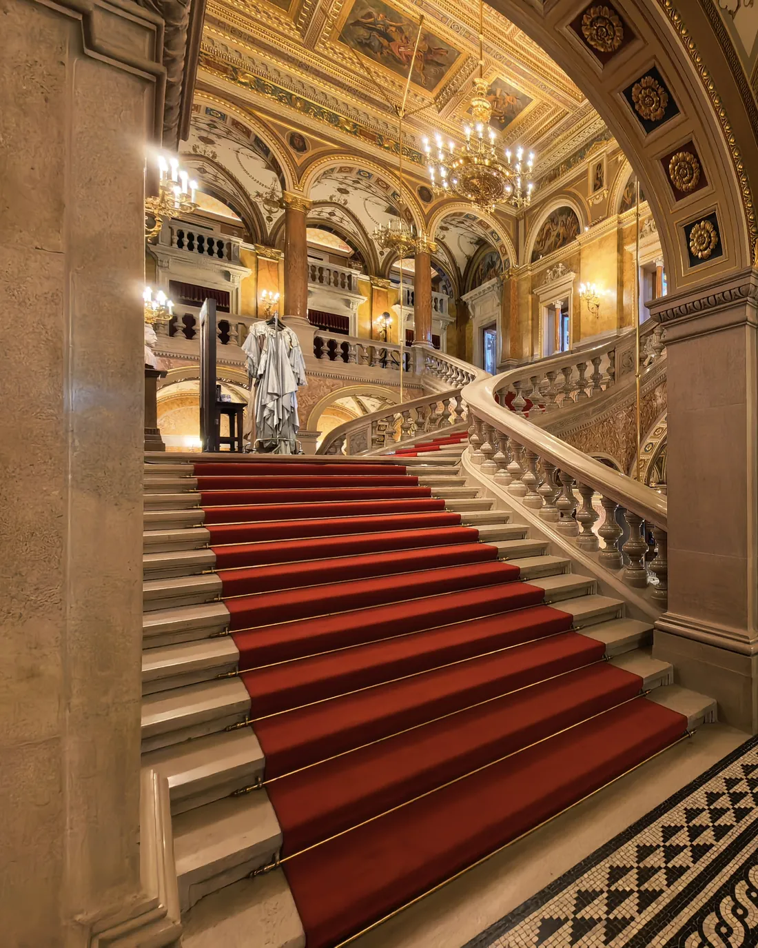 Una escalera de piedra con alfombra roja y balaustrada tallada que asciende bajo bóvedas pintadas en el interior de la Ópera
